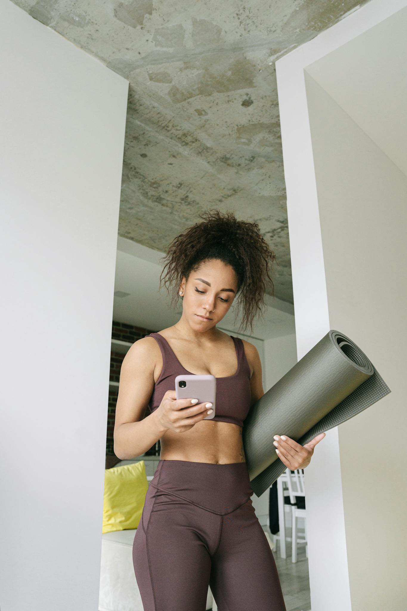 Fit young woman holding a yoga mat while looking at her phone indoors, ready for a workout.